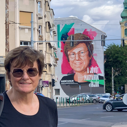 Katalin Kariko stands next to a mural of her portrait on the side of a building